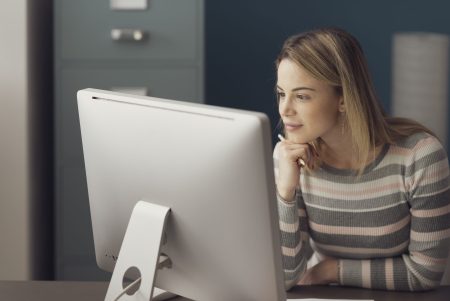Young attractive woman sitting at desk and working with her computer, she is smiling and looking at the screen
